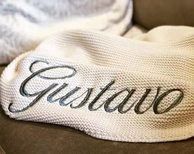 Cobija tejida de color crema con el nombre «Gustavo» bordado en letra cursiva gris.
