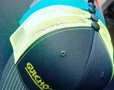 Gorra trucker negra con malla amarillo fluorescente y turquesa, con un logotipo bordado en hilo verde neón sobre la visera frontal.