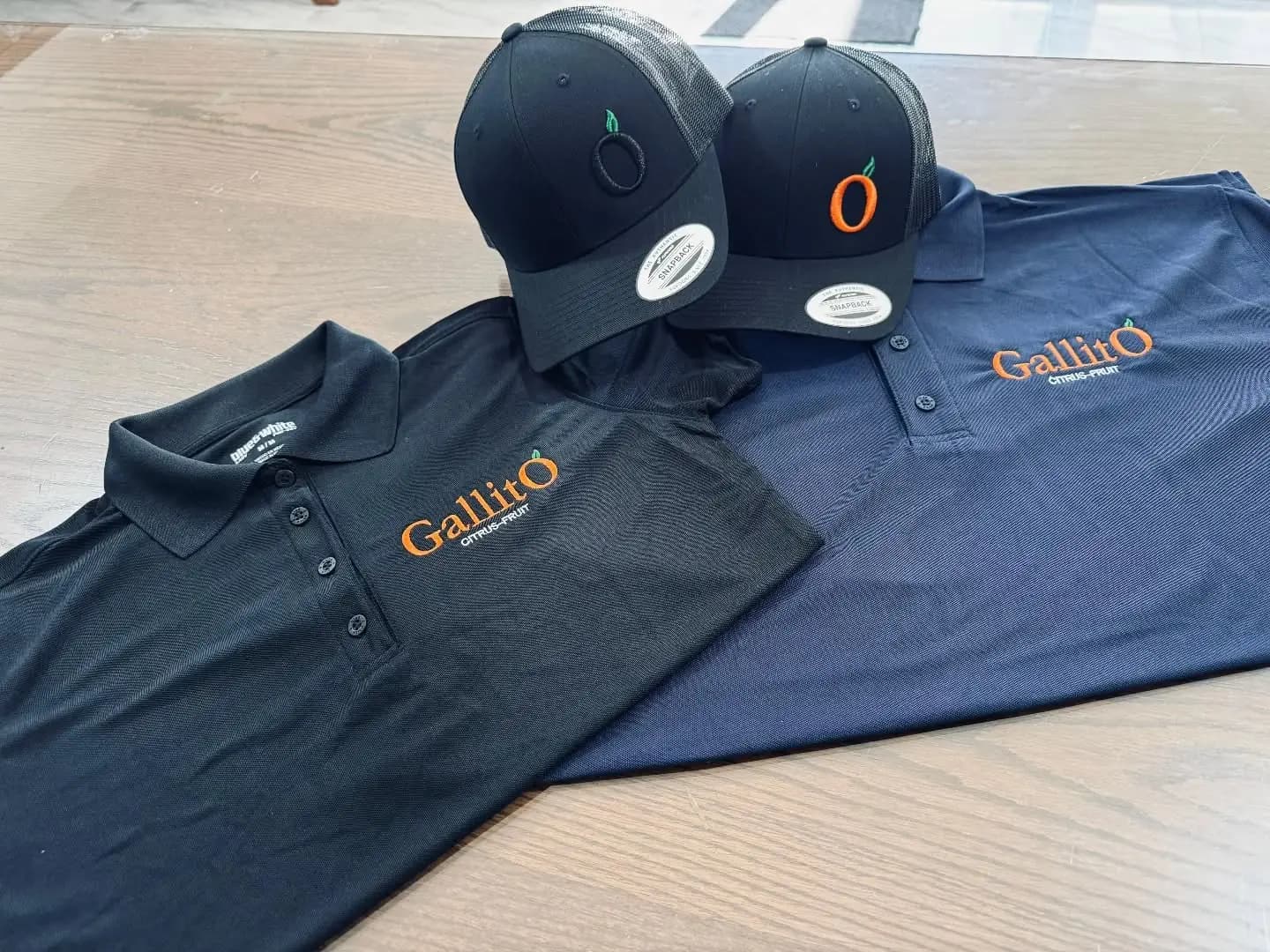 Dos playeras tipo polo, negra y azul marino, junto a dos gorras negras; todas con el logotipo bordado en naranja de Gallito Citrus-Fruit.