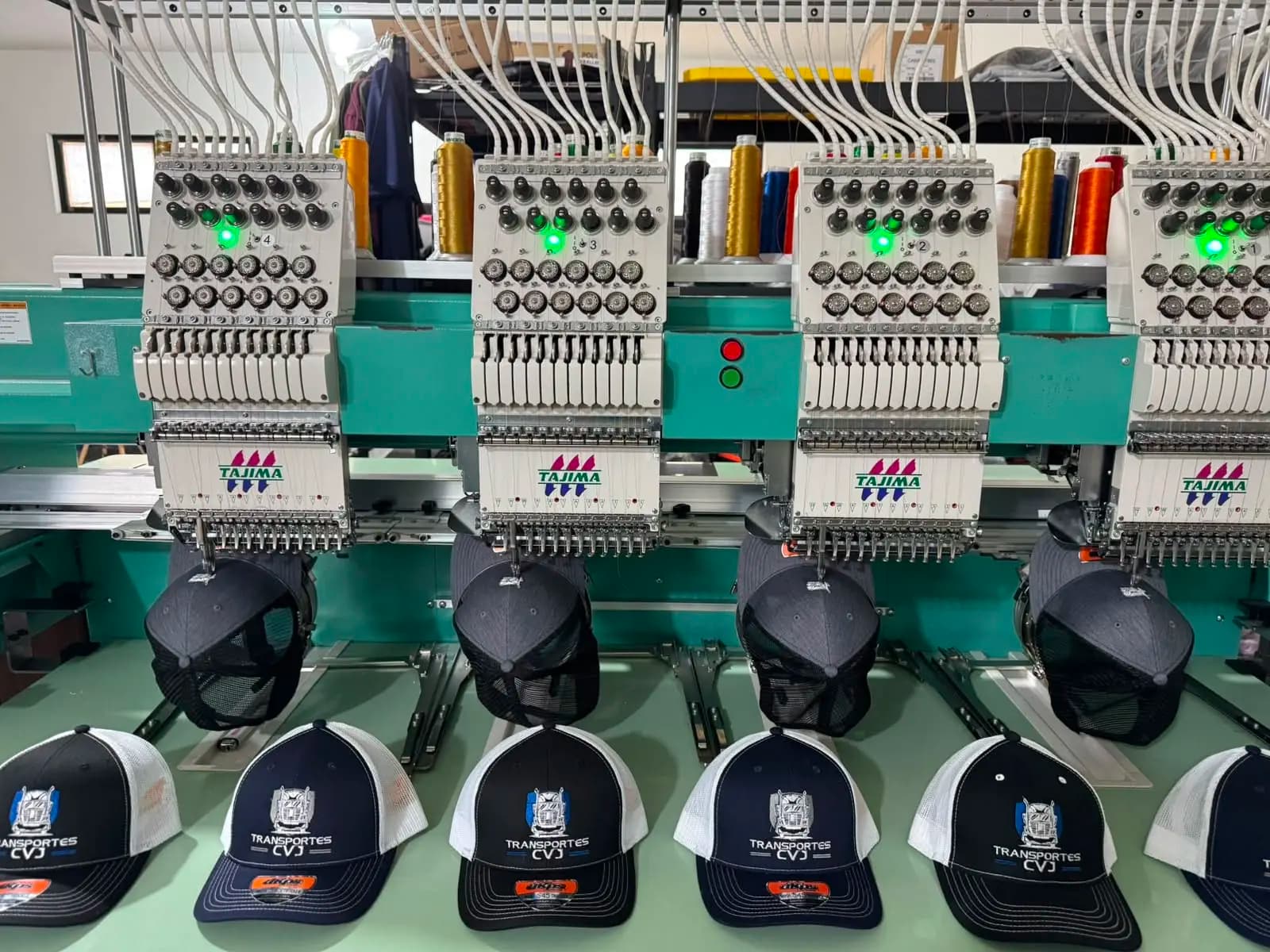 Máquina de bordado Tajima de cuatro cabezales trabajando sobre gorras colocadas en sus bastidores, con los conos de hilo de colores arriba y gorras ya terminadas de Transportes CVJ en primer plano.
