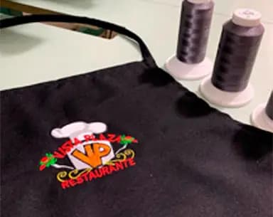 Mandil negro con un logotipo bordado a varios colores de un restaurante, junto a dos conos de hilo gris sobre la mesa de trabajo.